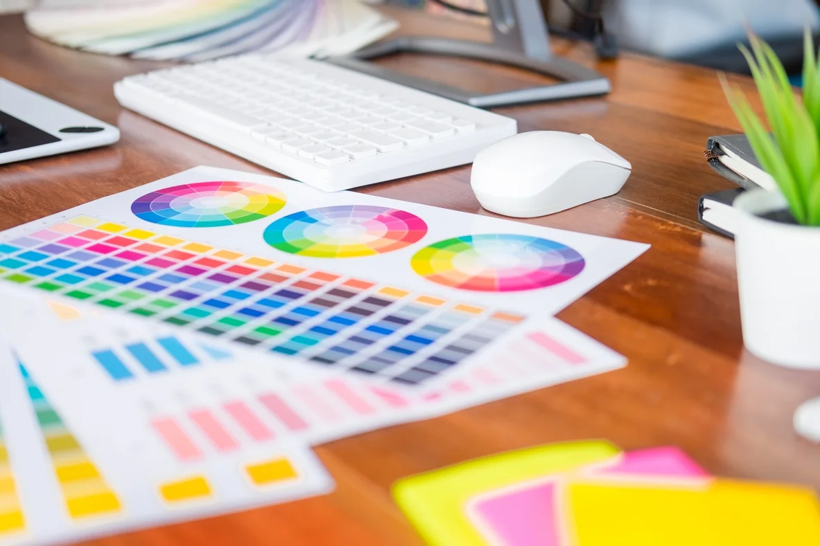 Un bureau de designer encombré avec , une roue chromatique, des carnet, une plante une souris et un clavier